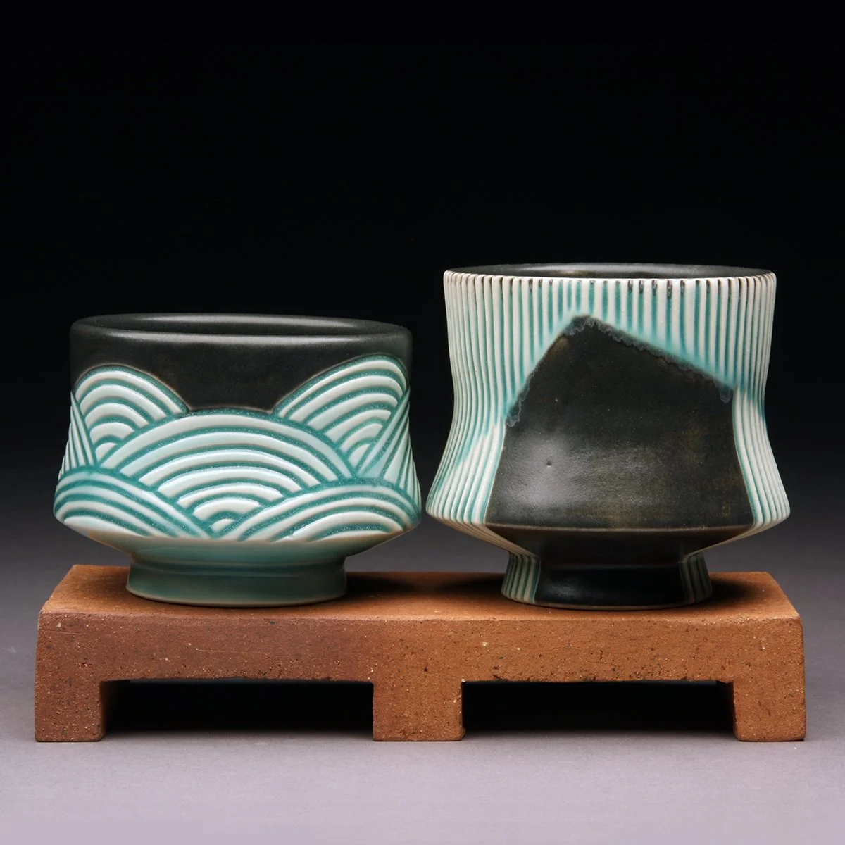 Two black and white ceramic cups with green wave patterns, placed on a small wooden stand against a black background.
