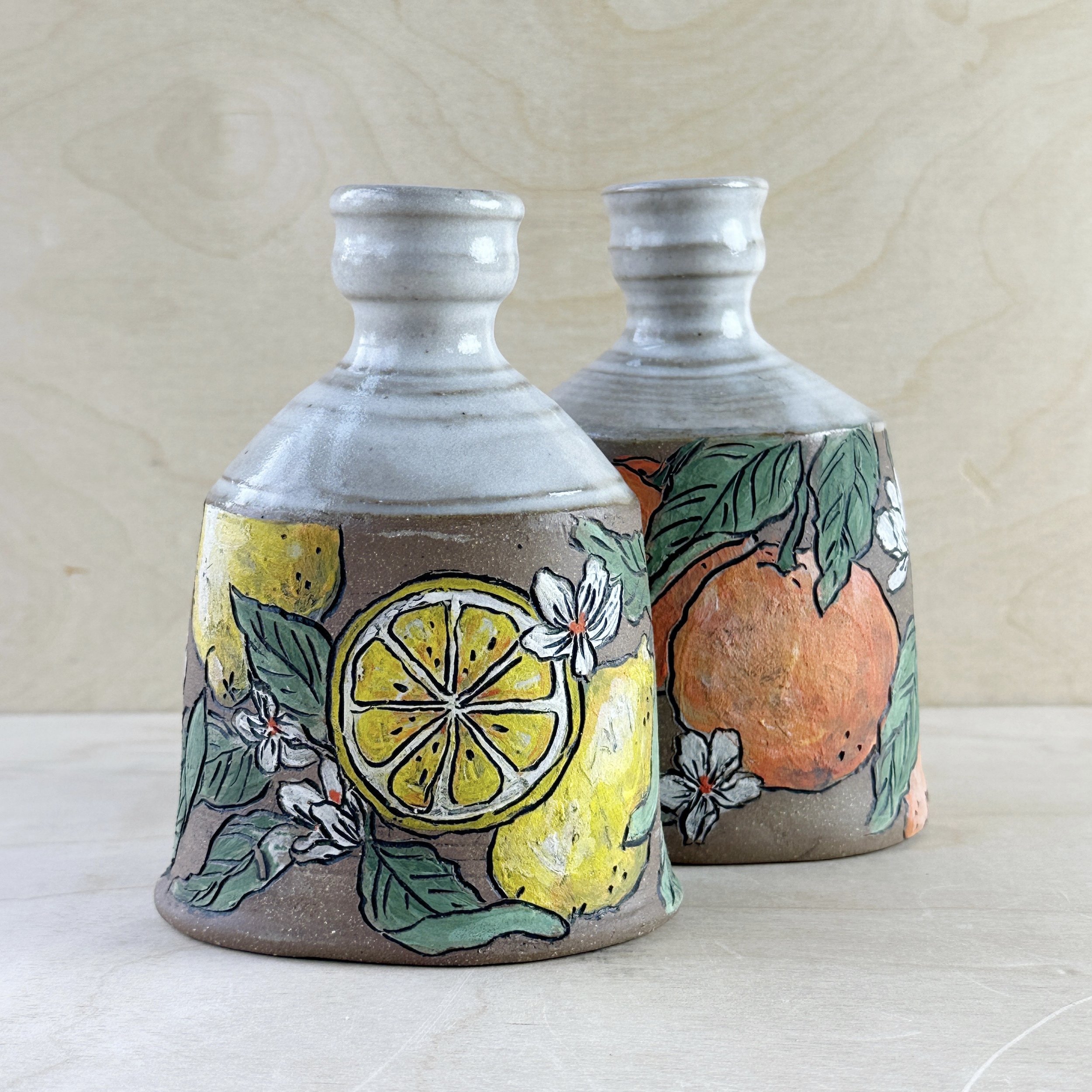 Two ceramic vases with painted fruit and floral designs, including lemons and oranges, on a wooden background.