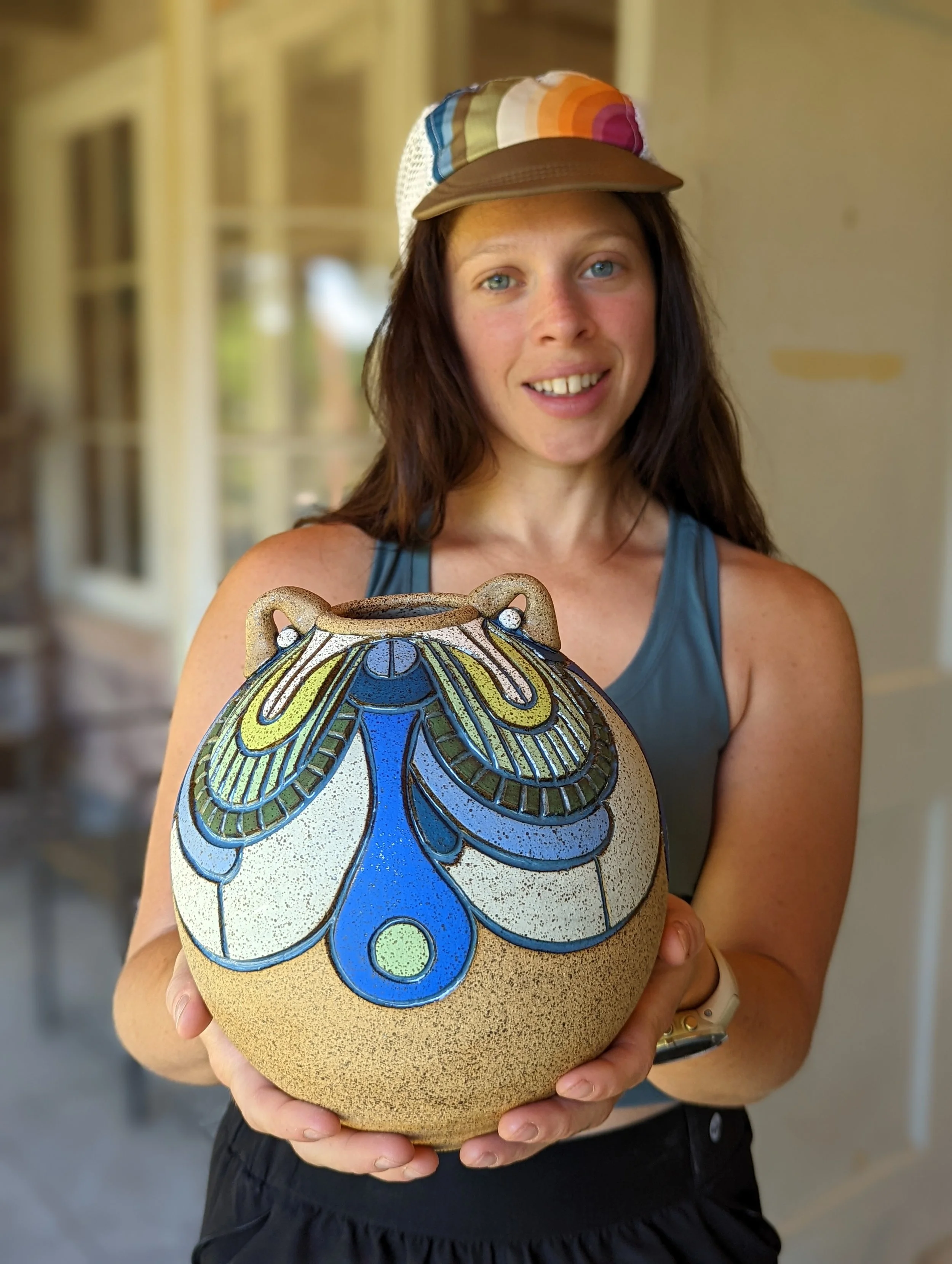 A woman with long brown hair wearing a multicolored hat and blue tank top holding a decorative ceramic vase with butterfly and wing designs.
