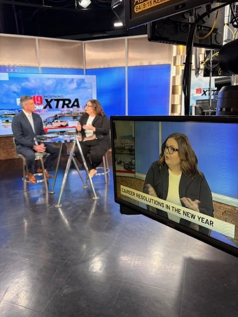 A television news studio with two hosts and a large monitor showing a woman, with a caption reading 'Career Resolutions in the New Year'.