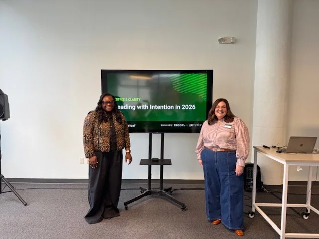 Two women standing in a conference room next to a large screen displaying a presentation slide about leading with intention in 2026. One woman is wearing a leopard print jacket and black pants, and the other is wearing a pink striped shirt and blue jeans.