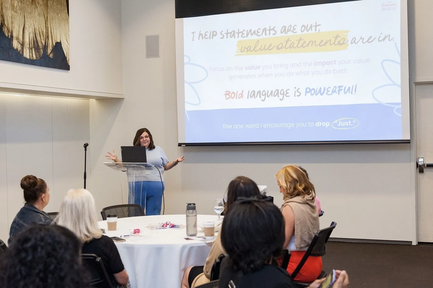 A woman giving a presentation in front of a seated audience in a conference room. The presentation slide behind her has text about statement impact and bold language.