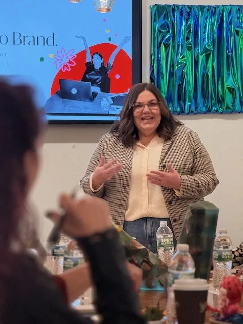 A woman giving a presentation in a room with a large screen behind her, displaying a graphic with the text 'Brand.' She is smiling, wearing glasses, a beige blazer, and a cream-colored shirt. There are tables with water bottles, snacks, and a green tumbler in the foreground.