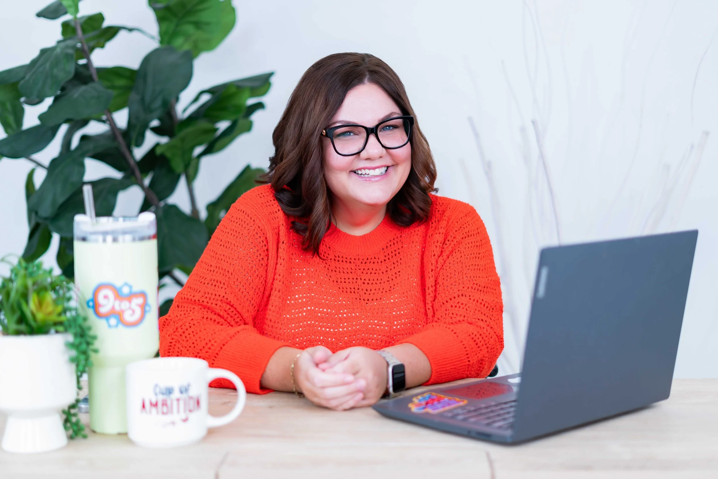 A woman with shoulder-length brown hair, wearing glasses and an orange sweater, sitting at a wooden table with a laptop, mugs, a plant, and a tumbler, smiling at the camera.