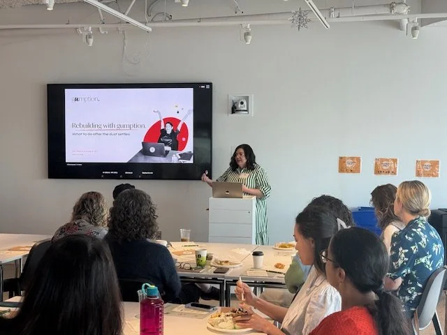 Woman giving a presentation in a conference room with seated attendees, a large screen displaying a slide that reads 'Rebuilding with Gumption' and a graphic of a woman at a desk.