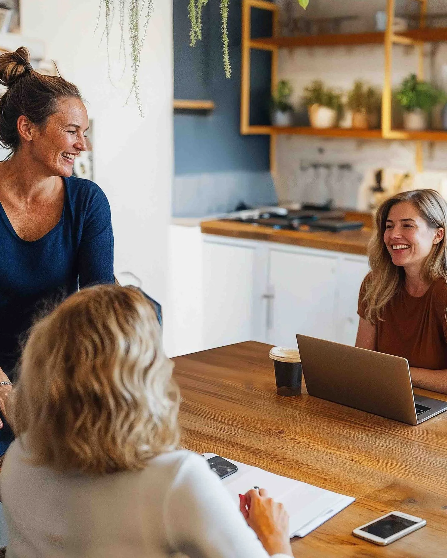 Drei Frauen in einem modernen Büro oder Café sitzen an einem Holztisch. Zwei Frauen lachen, während die dritte den Rücken zur Kamera hat. Ein Laptop, ein Smartphone und eine Kaffeetasse befinden sich auf dem Tisch.