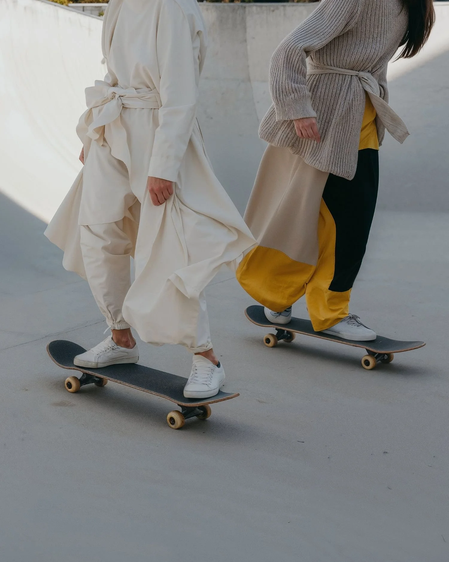 Zwei Personen mit fließenden, stilvollen Kleidungsstücken fahren auf Skateboards.
