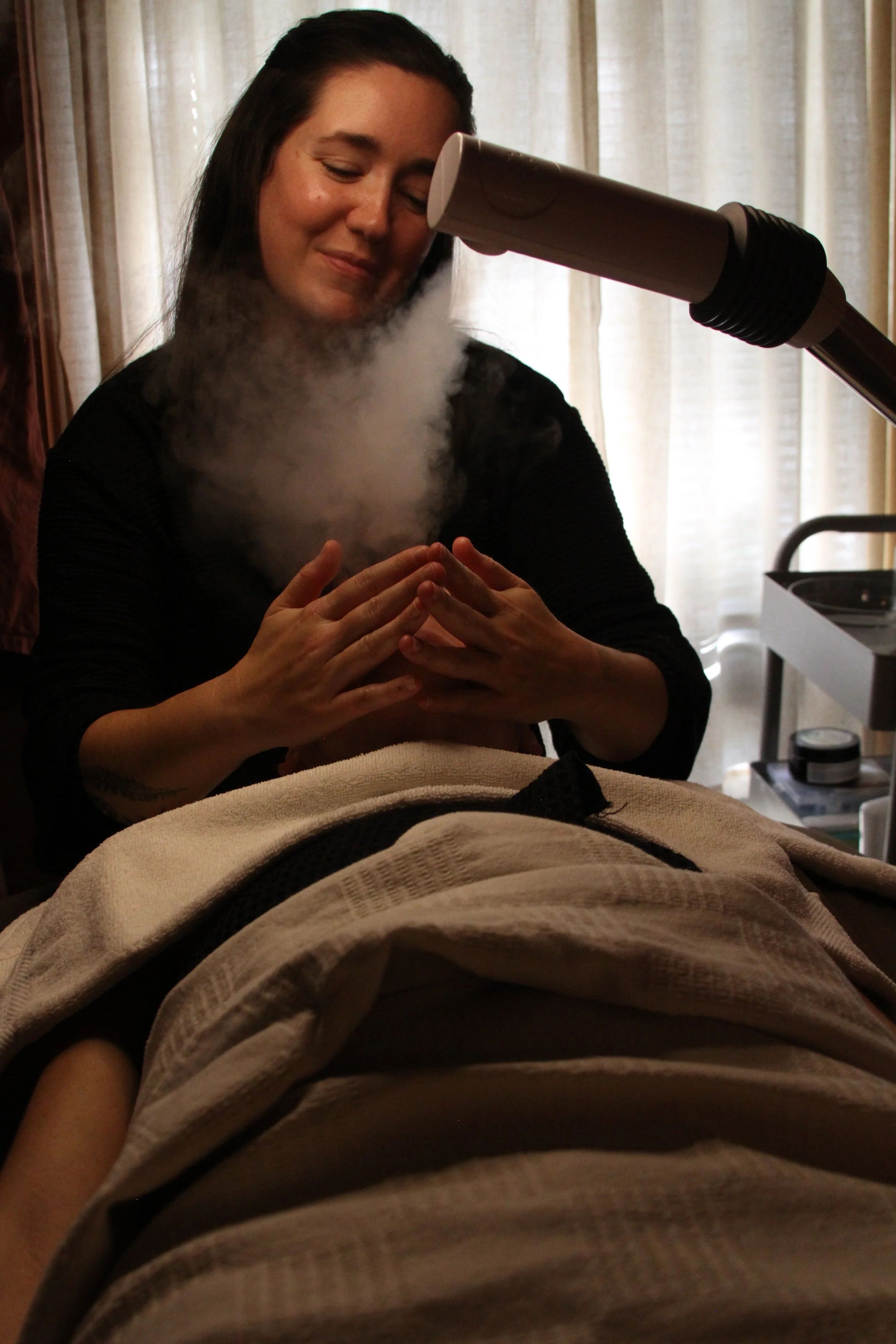 A woman receiving a steam facial treatment at a spa, with a technician using a steam device directed at her face, sitting in a treatment room with natural light.