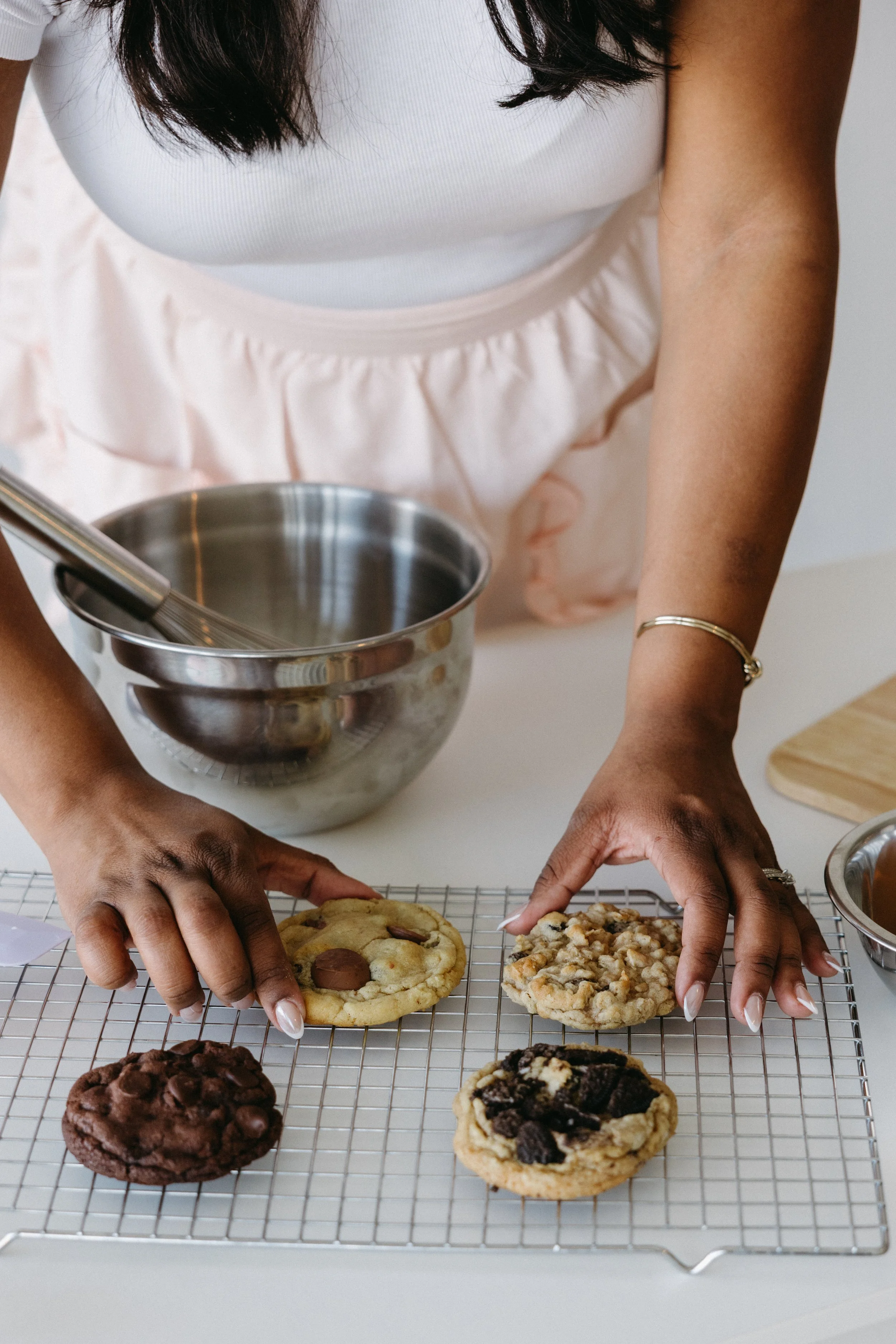 A person in a white top and pink apron arranging cookies on a cooling rack in a kitchen.