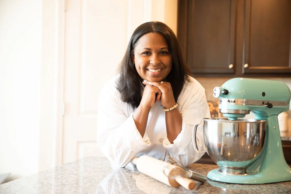 A woman smiling at the camera in a kitchen with a stand mixer, rolling pin, and dark cabinets.