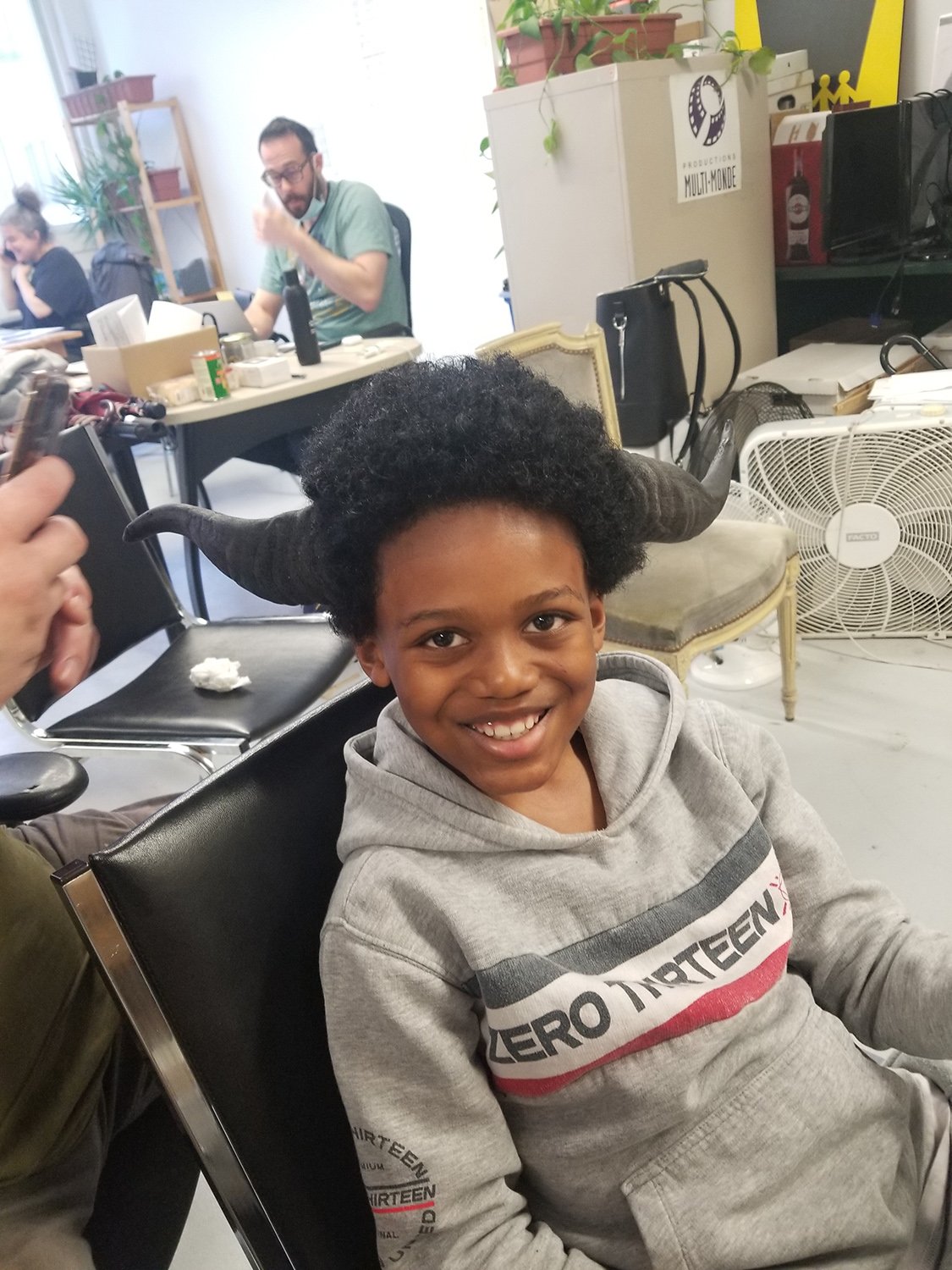 Enfant souriant portant un sweat-shirt "Zero Thirteen" avec des cornes d'animal attachées, assis dans un bureau avec des adultes travaillant en arrière-plan.