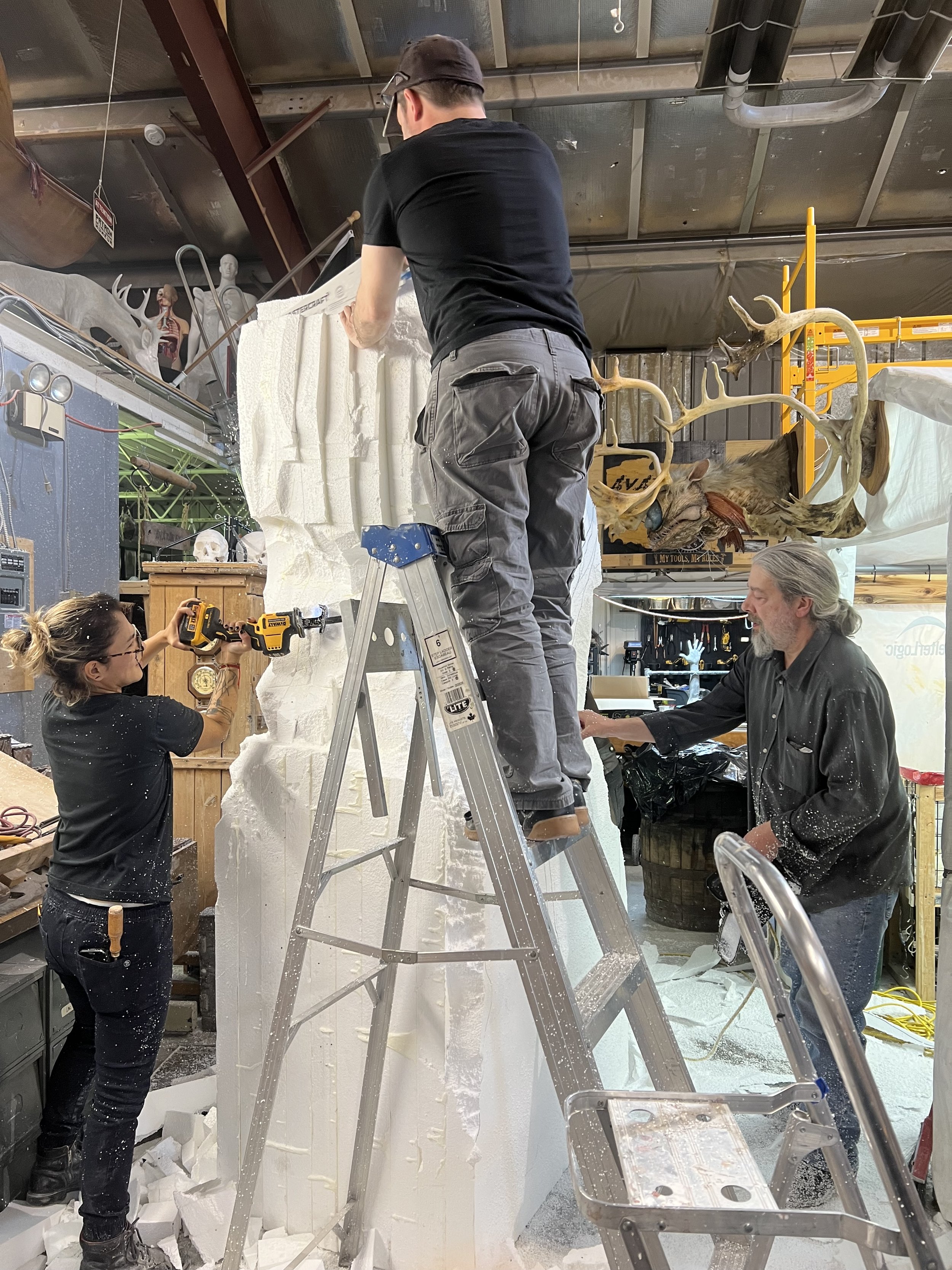 Trois personnes travaillant ensemble sur une grande sculpture en polystyrène dans un atelier, avec des outils et des échelles.