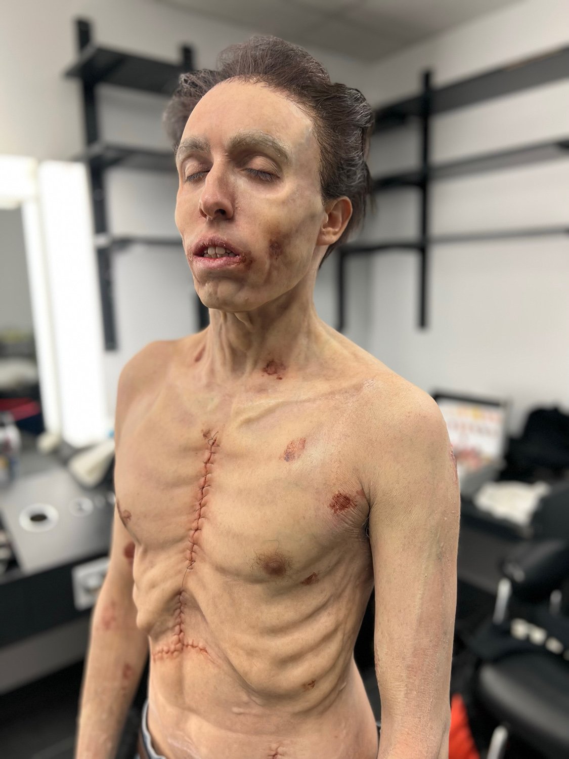 Personne avec maquillage d'effets spéciaux montrant un torse avec des cicatrices et des ecchymoses, dans une pièce avec des étagères en arrière-plan.