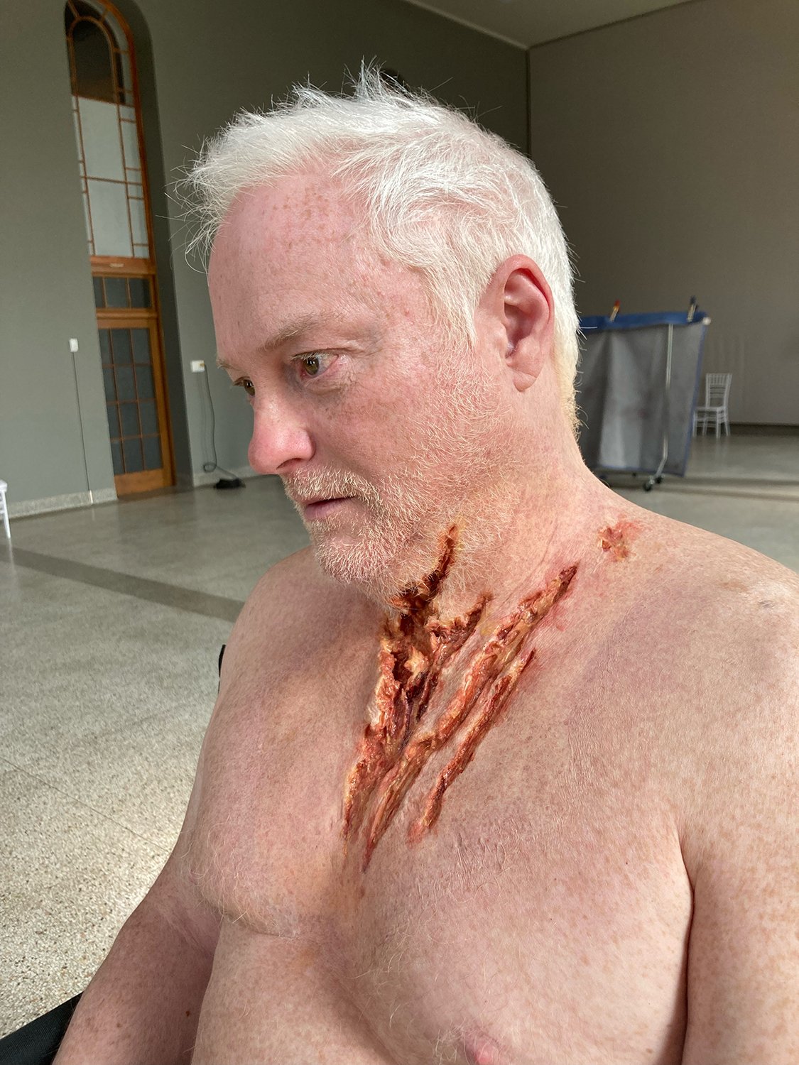 Un homme aux cheveux blancs avec un maquillage d'effets spéciaux simulant des blessures sur le cou et le torse, assis dans une pièce avec du mobilier en arrière-plan.