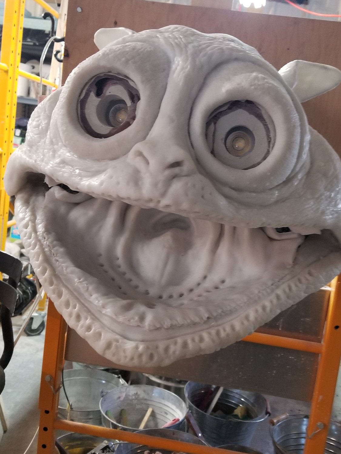 Sculpture inachevée d'une grande tête de créature avec des yeux proéminents et une bouche ouverte, posée sur une étagère orange dans un atelier.
