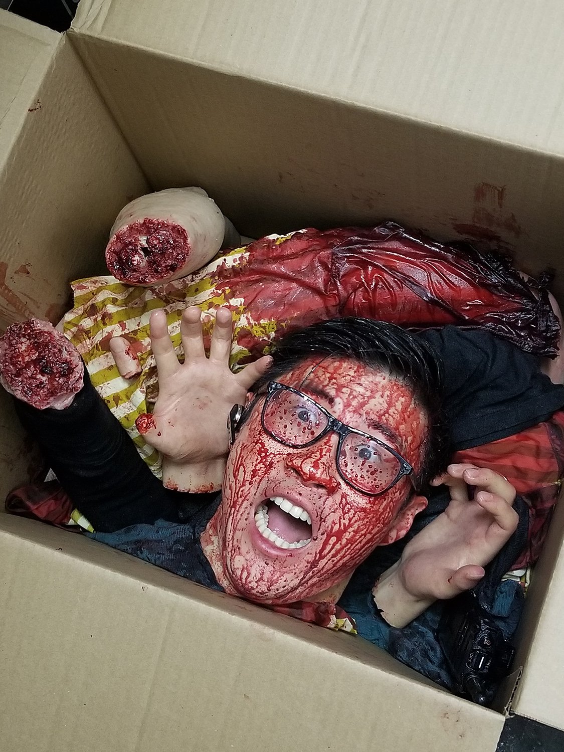 Une personne dans une boîte en carton avec du maquillage imitant du sang et des membres artificiels, semblant affolée.