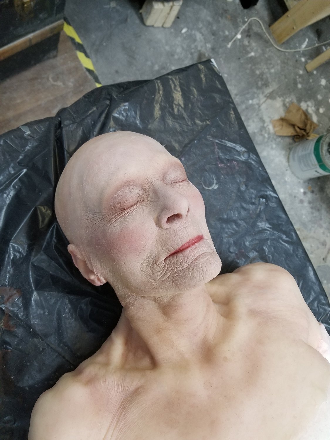 Mannequin imitant une personne âgée, allongé sur une surface noire, avec des détails réalistes du visage.