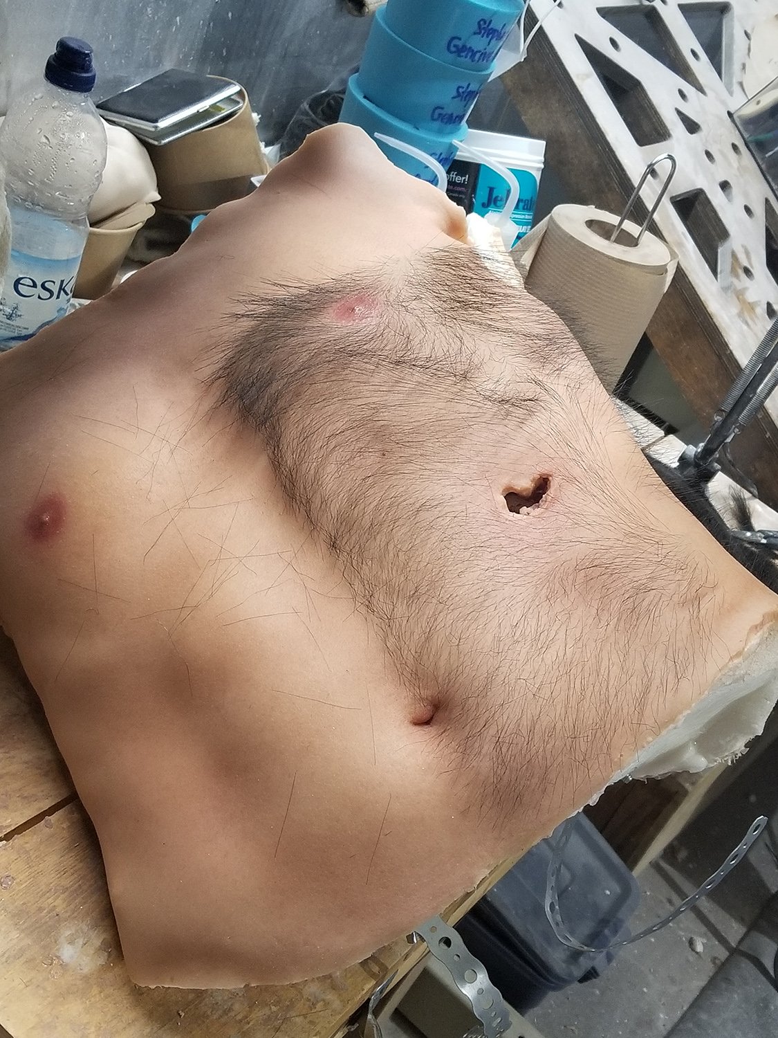 Un torse en silicone avec des poils, posé sur une table, entouré de bouteilles et autres objets.