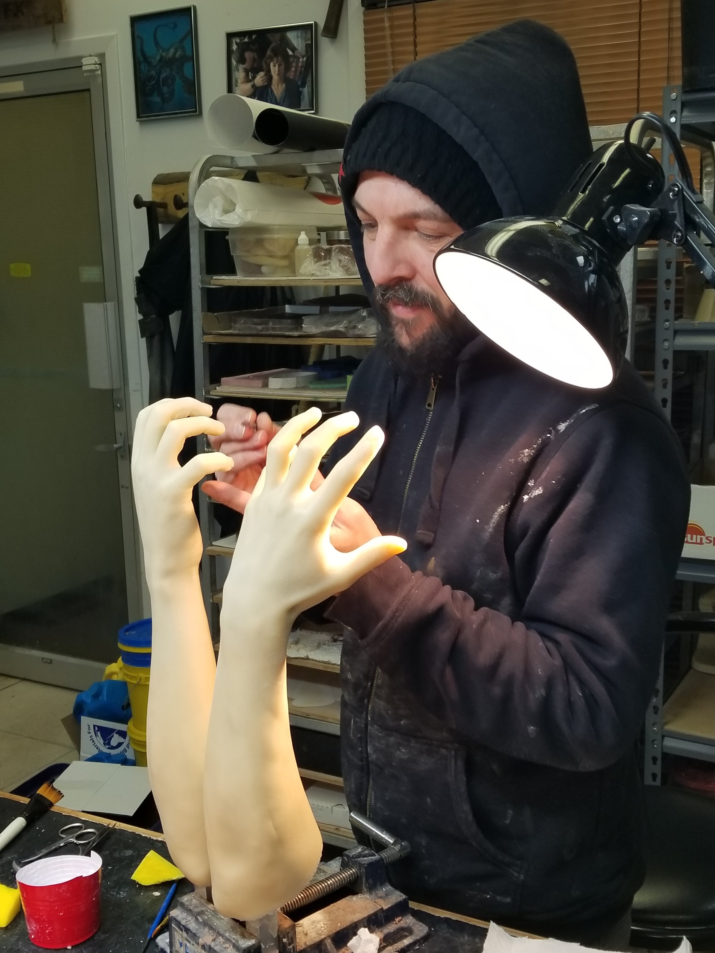 Une personne portant un bonnet, travaillant sur deux répliques réalistes de mains et avant-bras en utilisant des outils dans un atelier.
