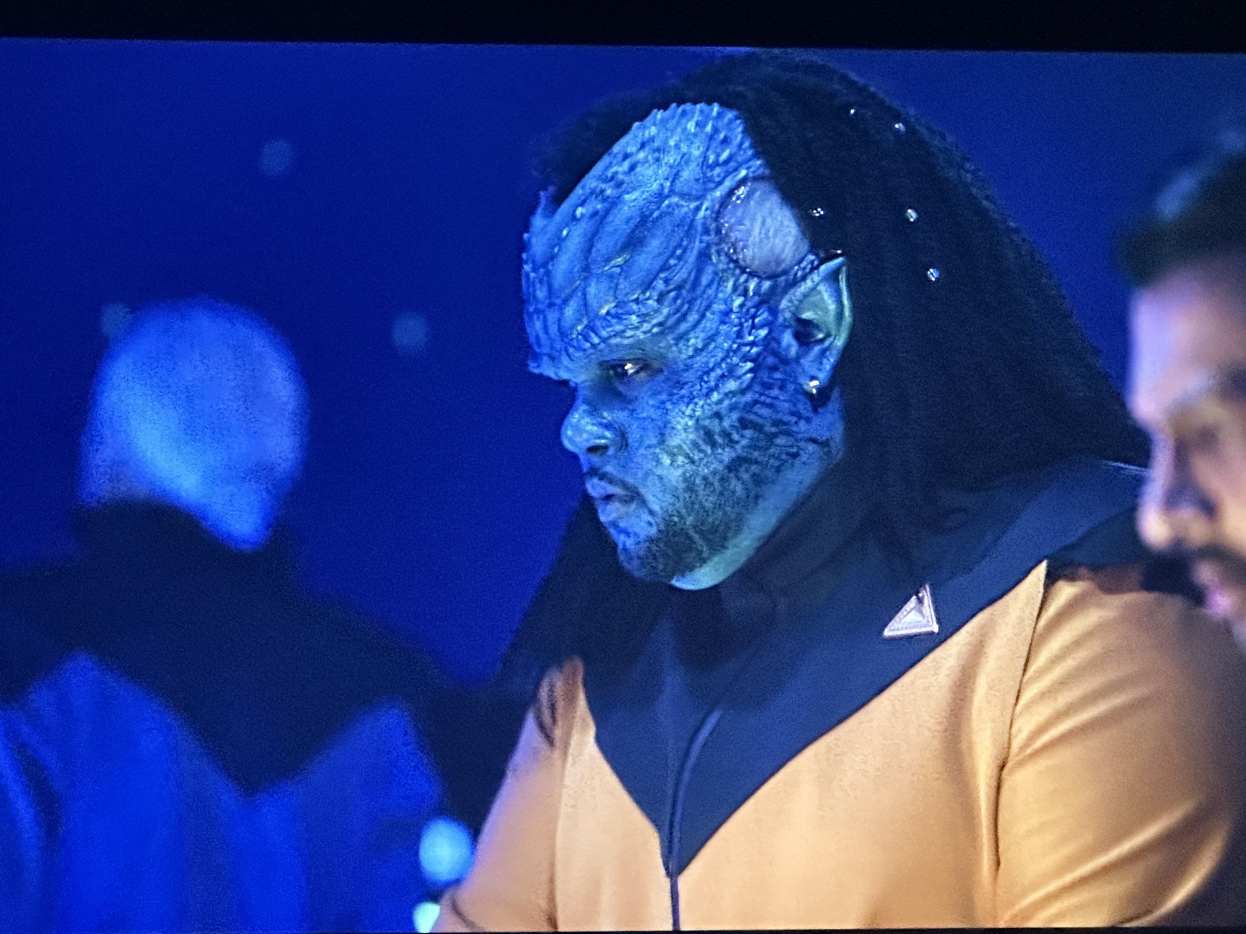 Personnage extraterrestre au visage bleu, habillé en tenue de science-fiction jaune et noire, fond bleu sombre.