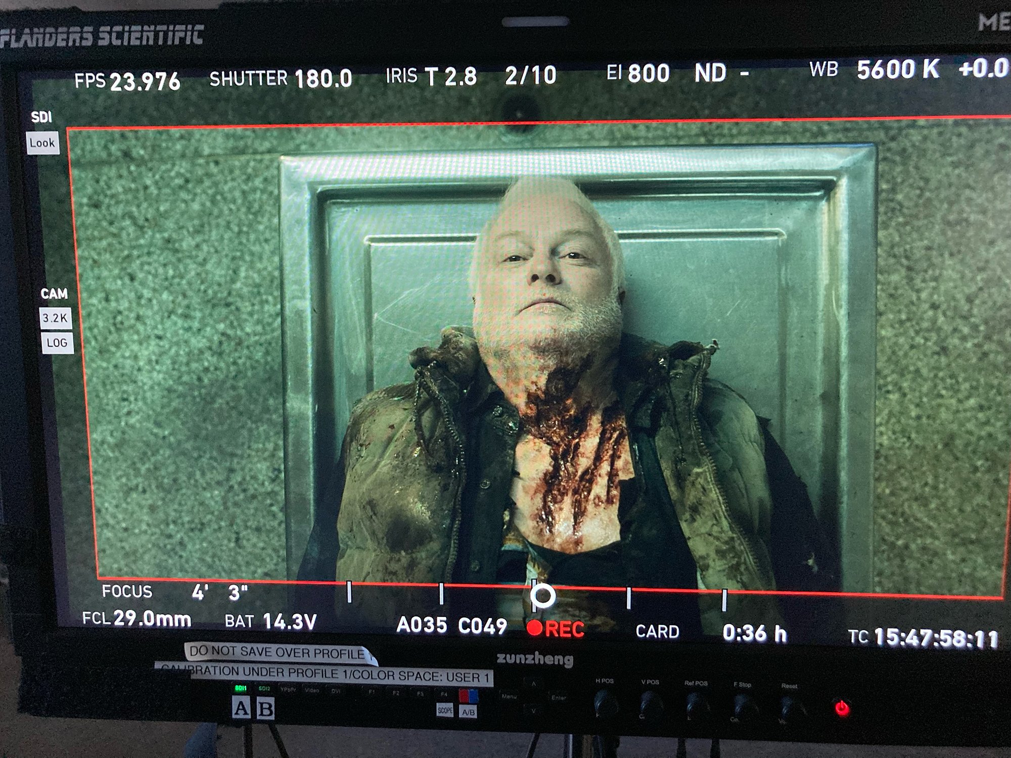 Moniteur de tournage montrant un homme allongé, portant des vêtements tachés de sang, sur un fond métallique.