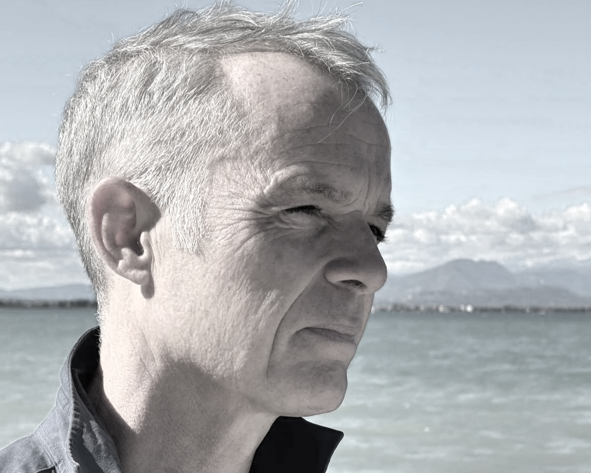 Close-up of a middle-aged man's face in profile, outdoors, with ocean and mountains in the background, black and white with some color.