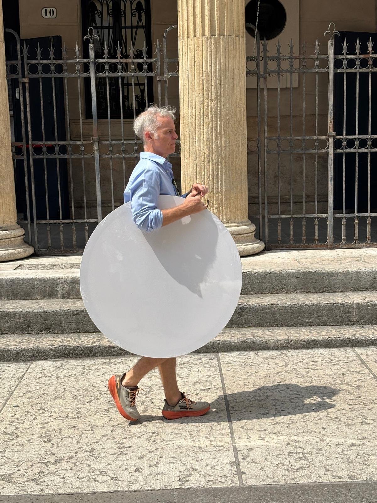 Man walking on sidewalk with a large, round, white reflective sign attached to his waist, wearing a light blue shirt, shorts, and sneakers, in front of a building with columns and iron gates.