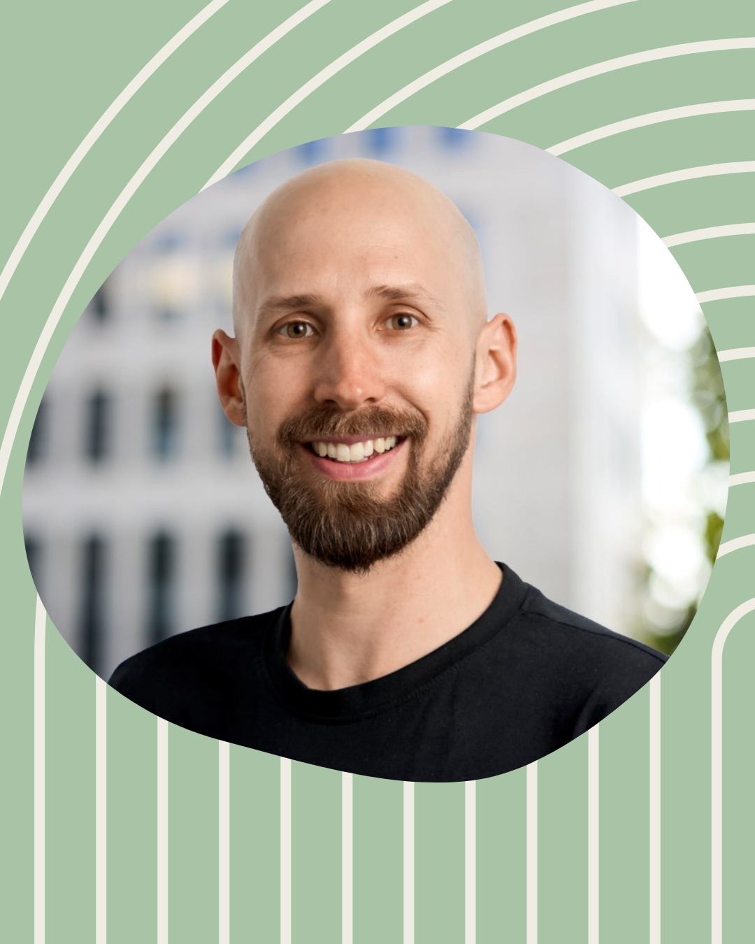 Ein lachender Mann mit einem Bart und einem schwarzen T-Shirt bei einer Stadtaufnahme im Hintergrund.