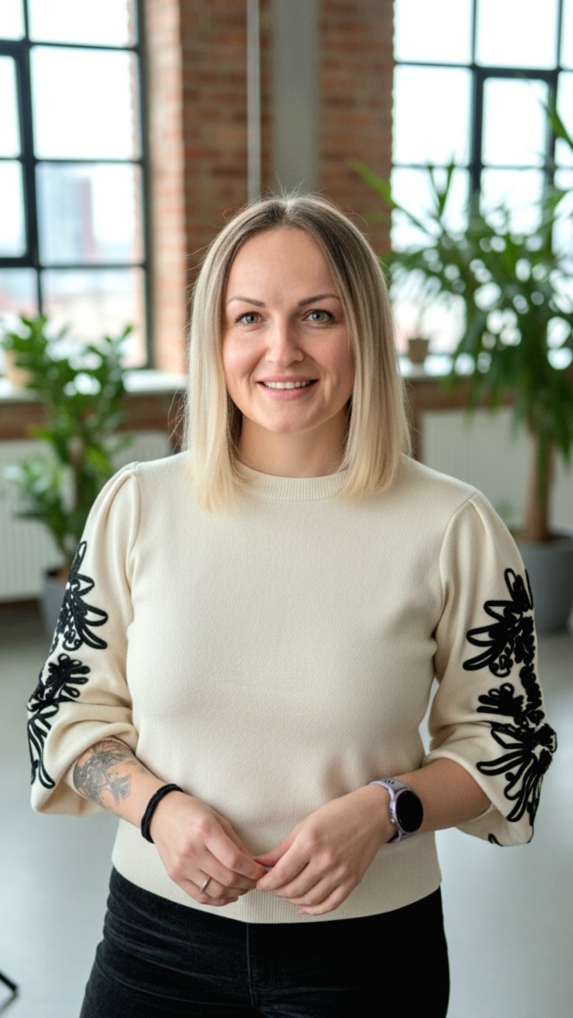 Eine junge Frau mit blonden, schulterlangen Haaren in einem hellen Pullover steht in einem modernen Raum mit großen Fenstern und grünen Pflanzen im Hintergrund, lächelt und blickt in die Kamera.