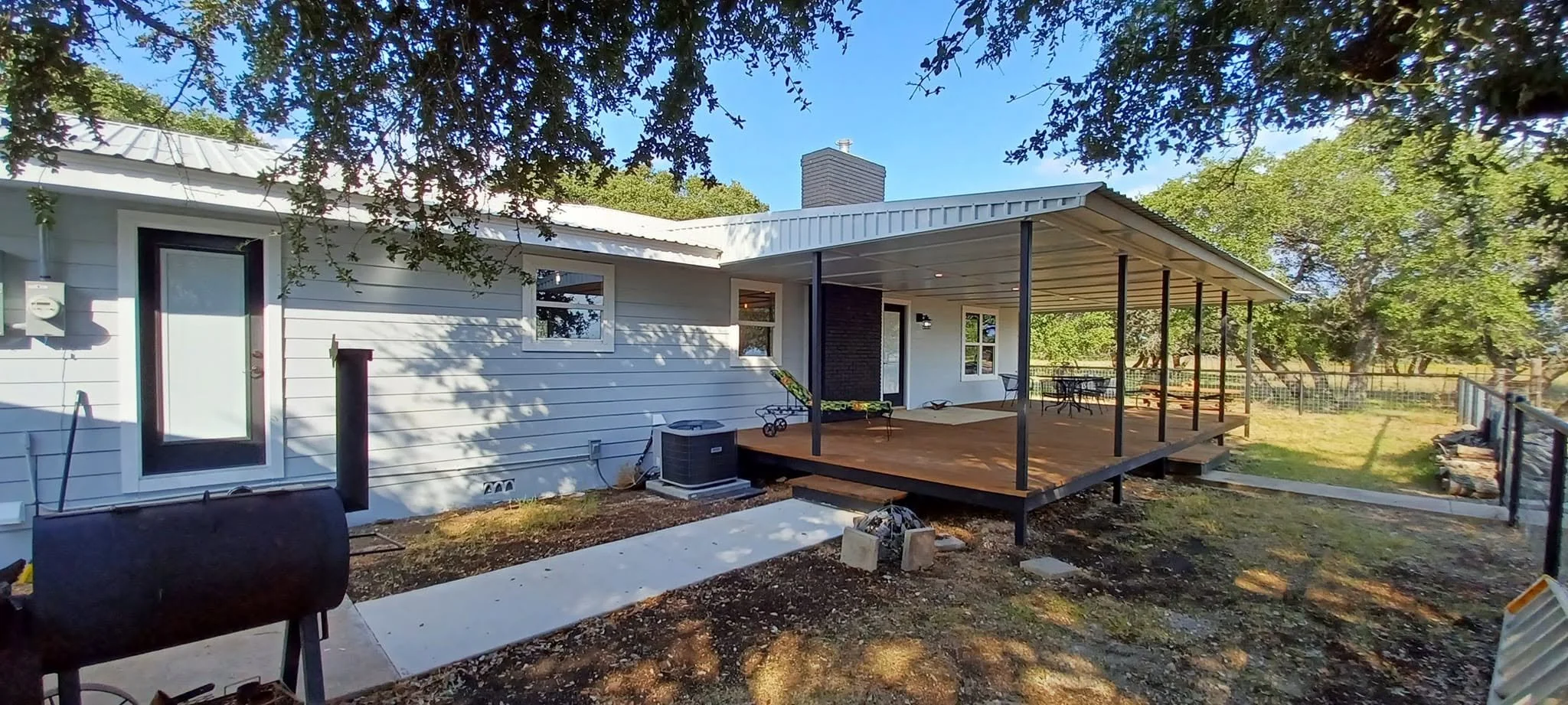 Backyard with a white house, wooden deck, outdoor table and chairs, surrounded by trees and fencing, during daytime.