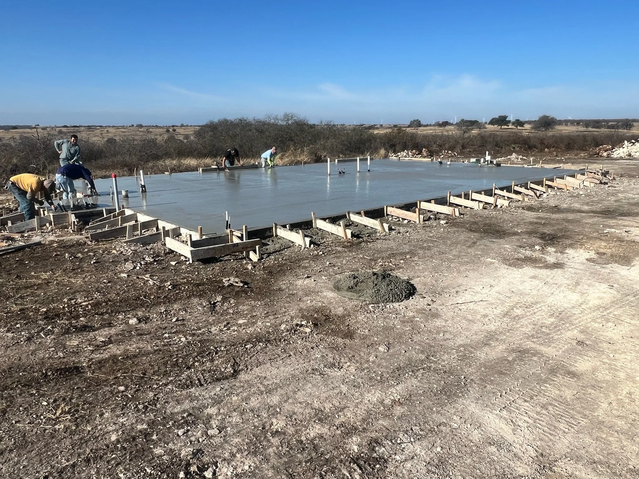People working on a concrete slab foundation at a construction site in a rural area with clear blue sky and wind turbines in the background.