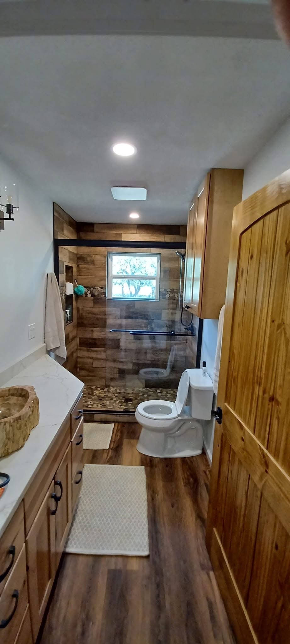A rustic bathroom with a wood-paneled shower area, a window, a toilet, and wooden cabinets, with hardwood floors and beige rugs.