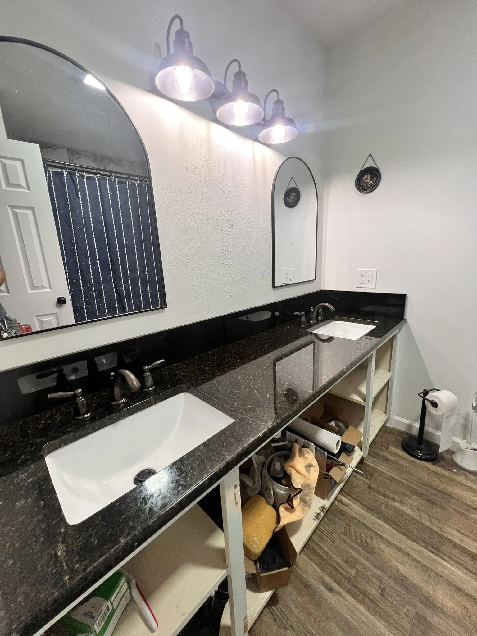 A bathroom with a black granite countertop, a white sink, a mirror, pendant lights, and open shelving underneath, with various tools and items stored on the shelves and floor.