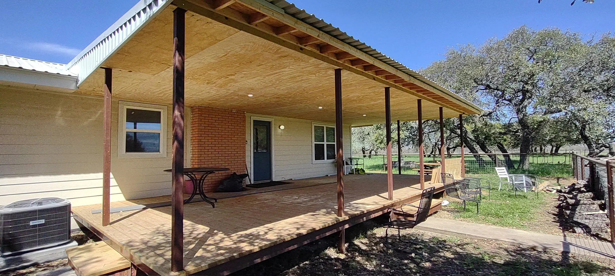 Back porch of a house with a new wooden deck under construction, supported by metal posts, surrounded by backyard with trees, outdoor chairs, and a wire fence.