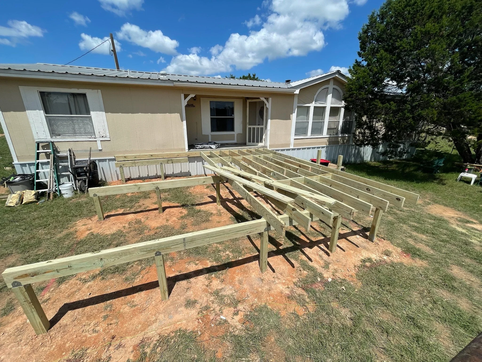 A house under construction with a frame for a deck or porch in front, construction materials and tools nearby, and an overgrown yard with trees and a blue sky with clouds.