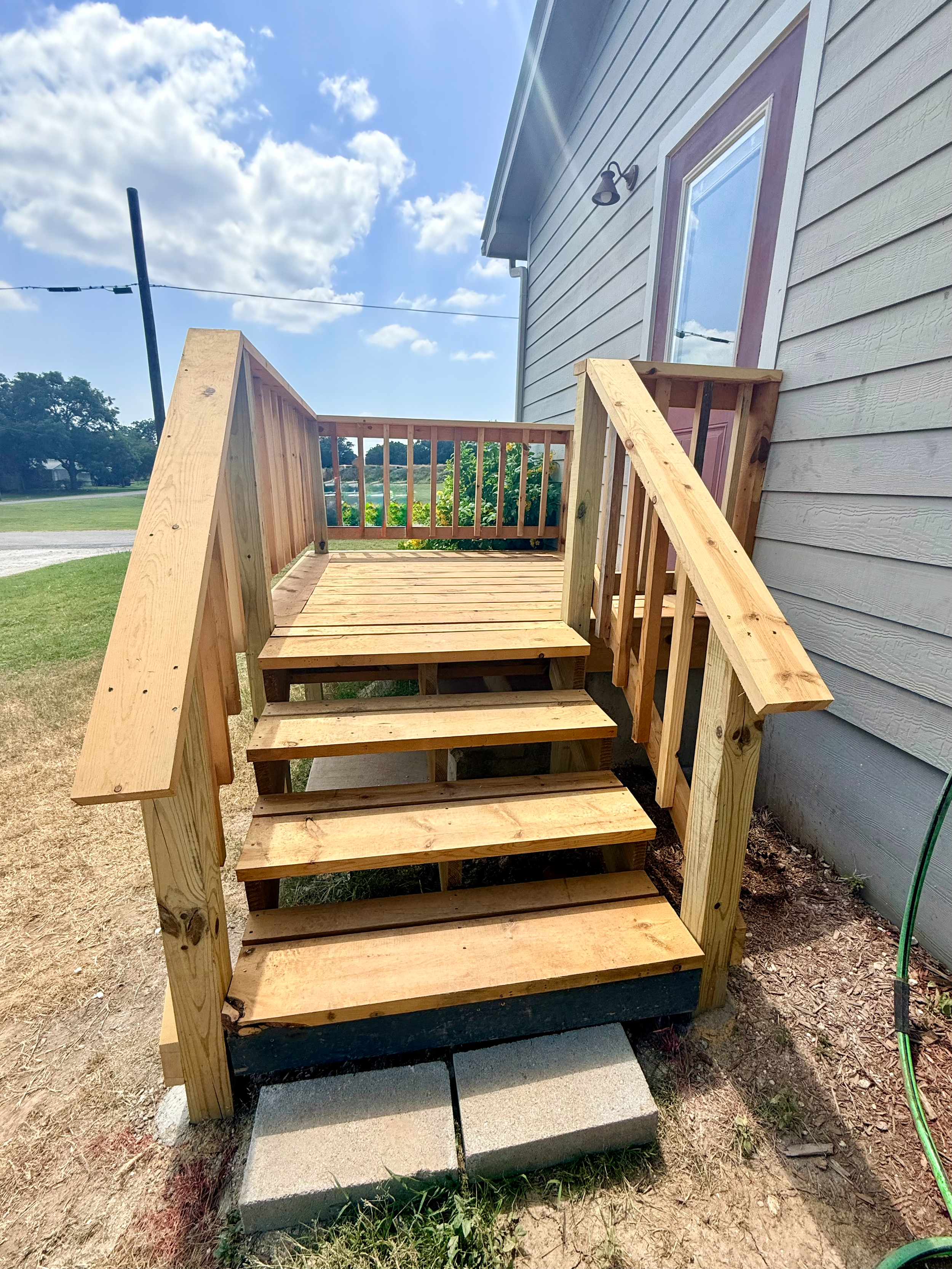A wooden staircase leading to a house door with a purple trim, outdoors on a sunny day, with a grassy yard, blue sky, and some clouds in the background.
