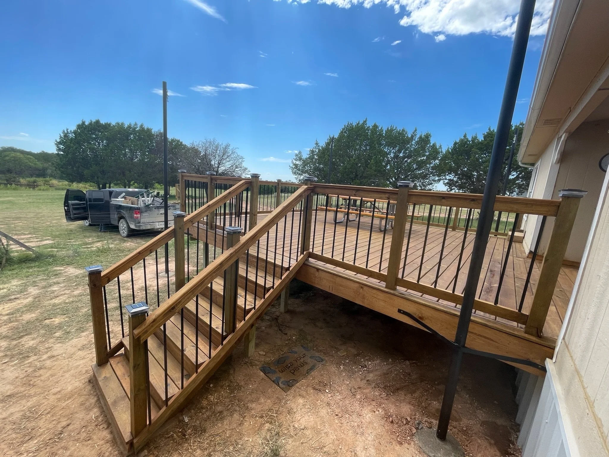 Backyard deck under construction with new wooden stairs and railings, a black pickup truck parked on a grassy area, trees, blue sky with some clouds, and part of a house with a clock and gutter