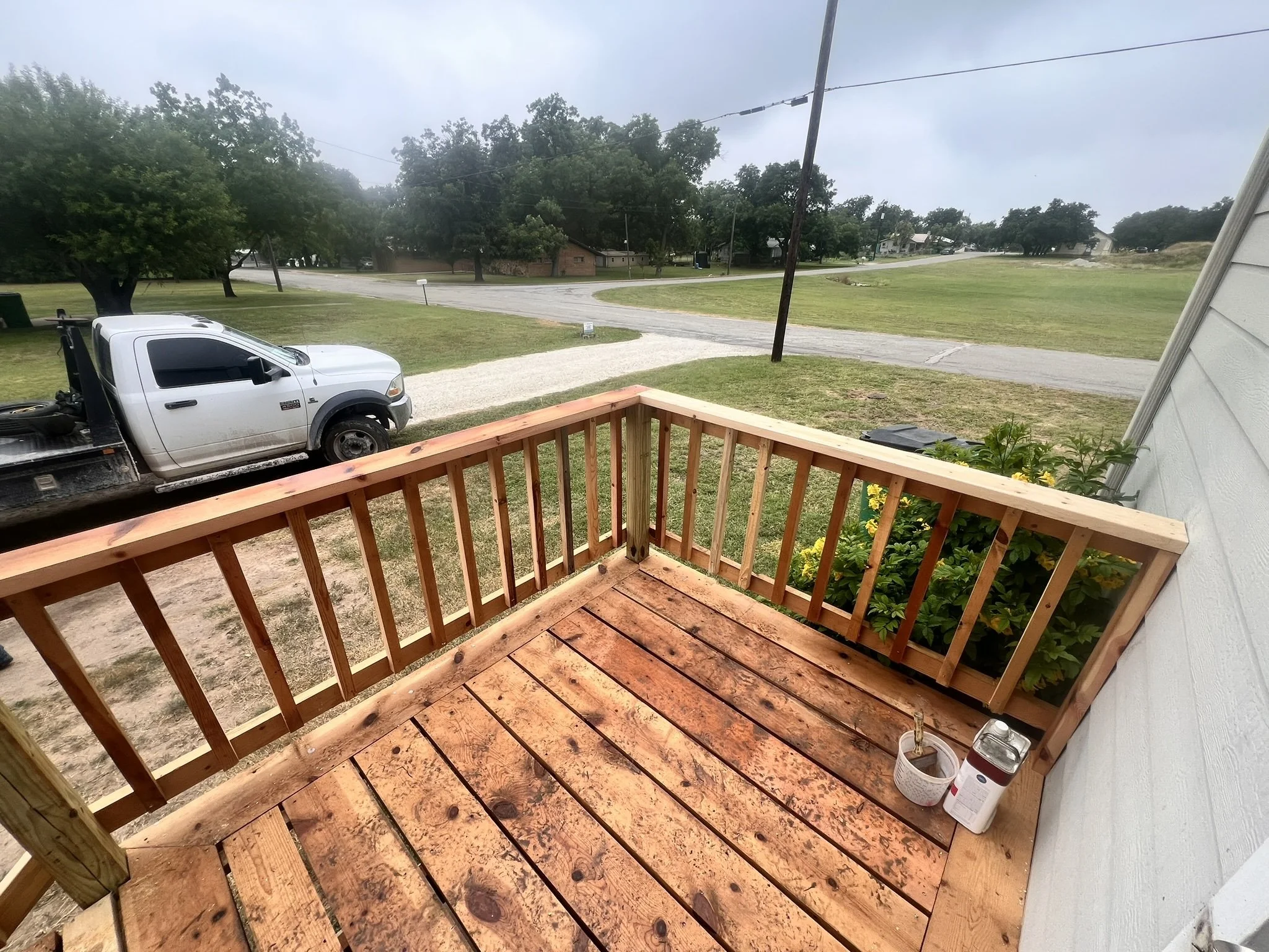View from a porch showing a wooden deck, flower pots, a grassy yard, a white truck, trees, and a cloudy sky.