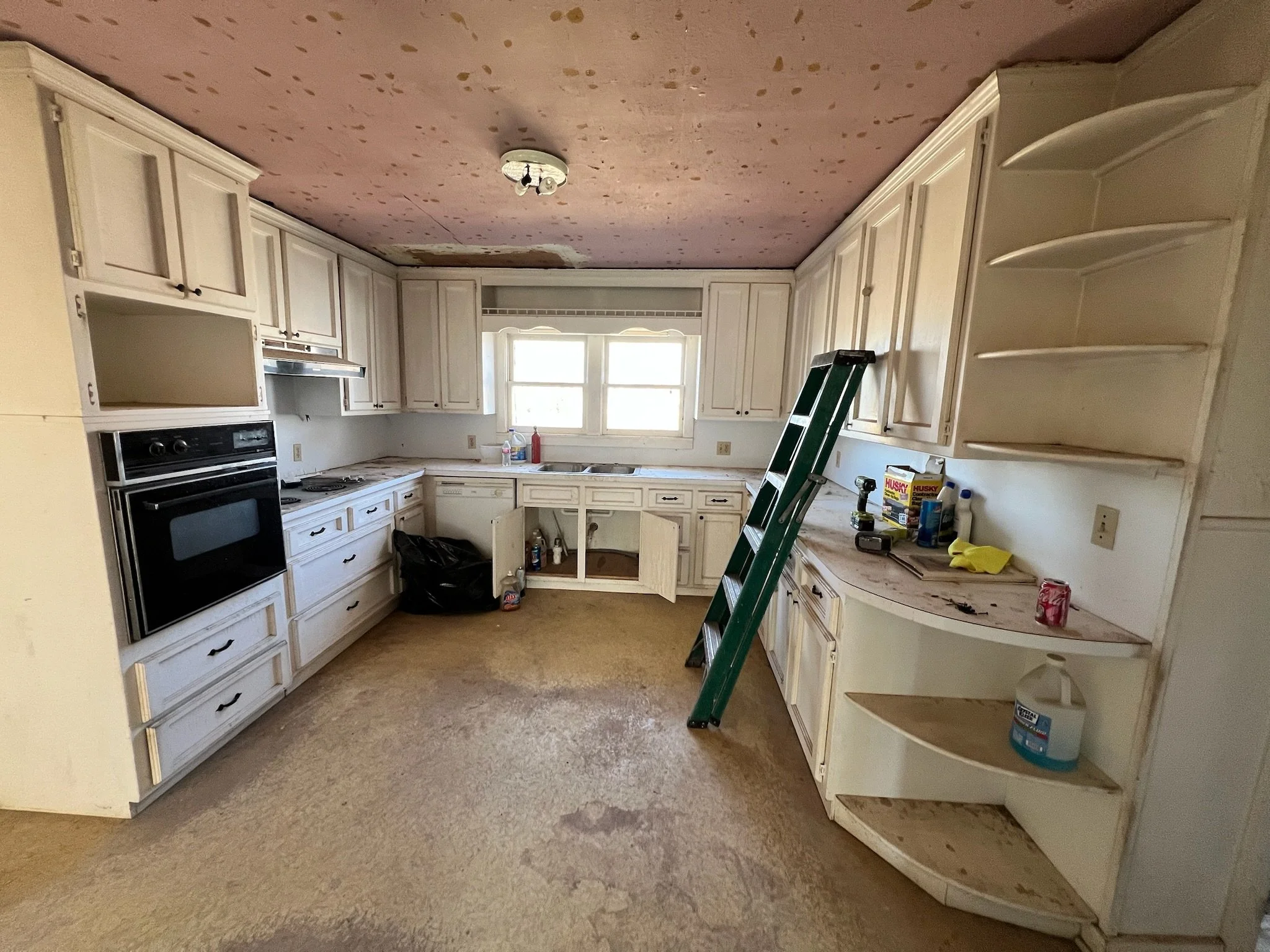 Kitchen with white cabinets, a window, and a partially assembled cabinet on the right. The ceiling has spots of paint or stain, and the floor appears worn. There are tools, cleaning supplies, and a ladder present.