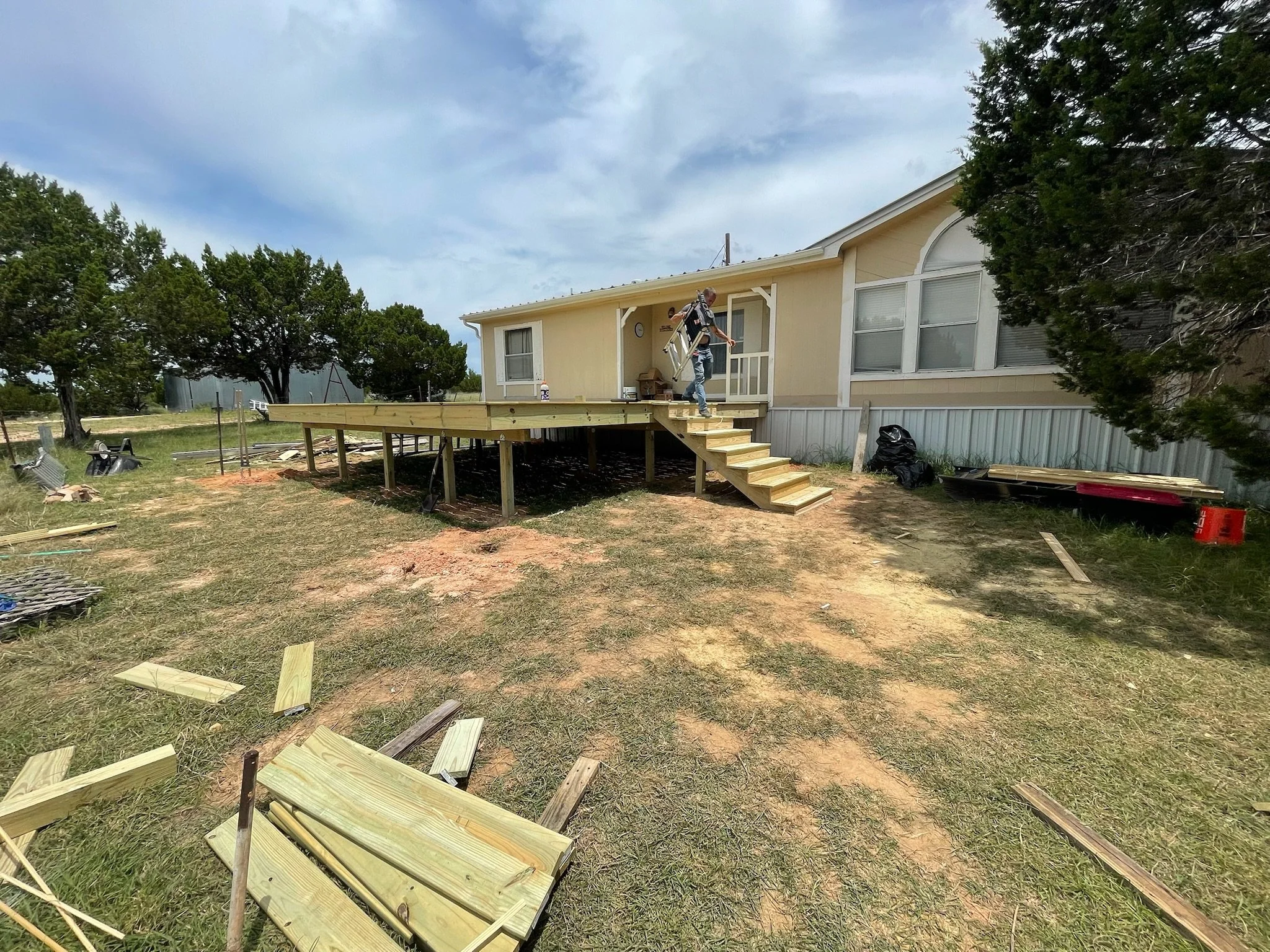 Person carrying a deck railing on a partially built wooden deck in the yard of a yellow house with trees and a cloudy sky in the background.