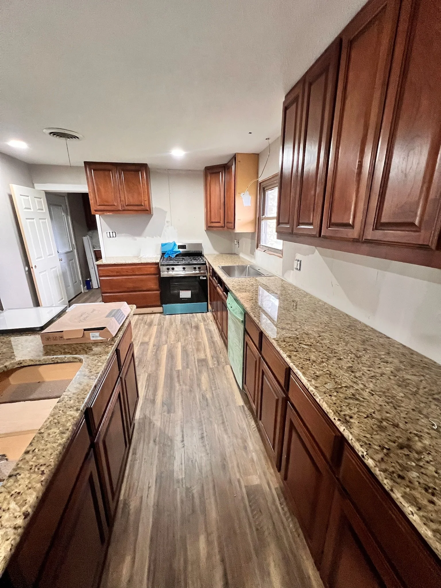 A kitchen under renovation with wooden cabinets, a granite countertop, a stove, a window, and wooden flooring.