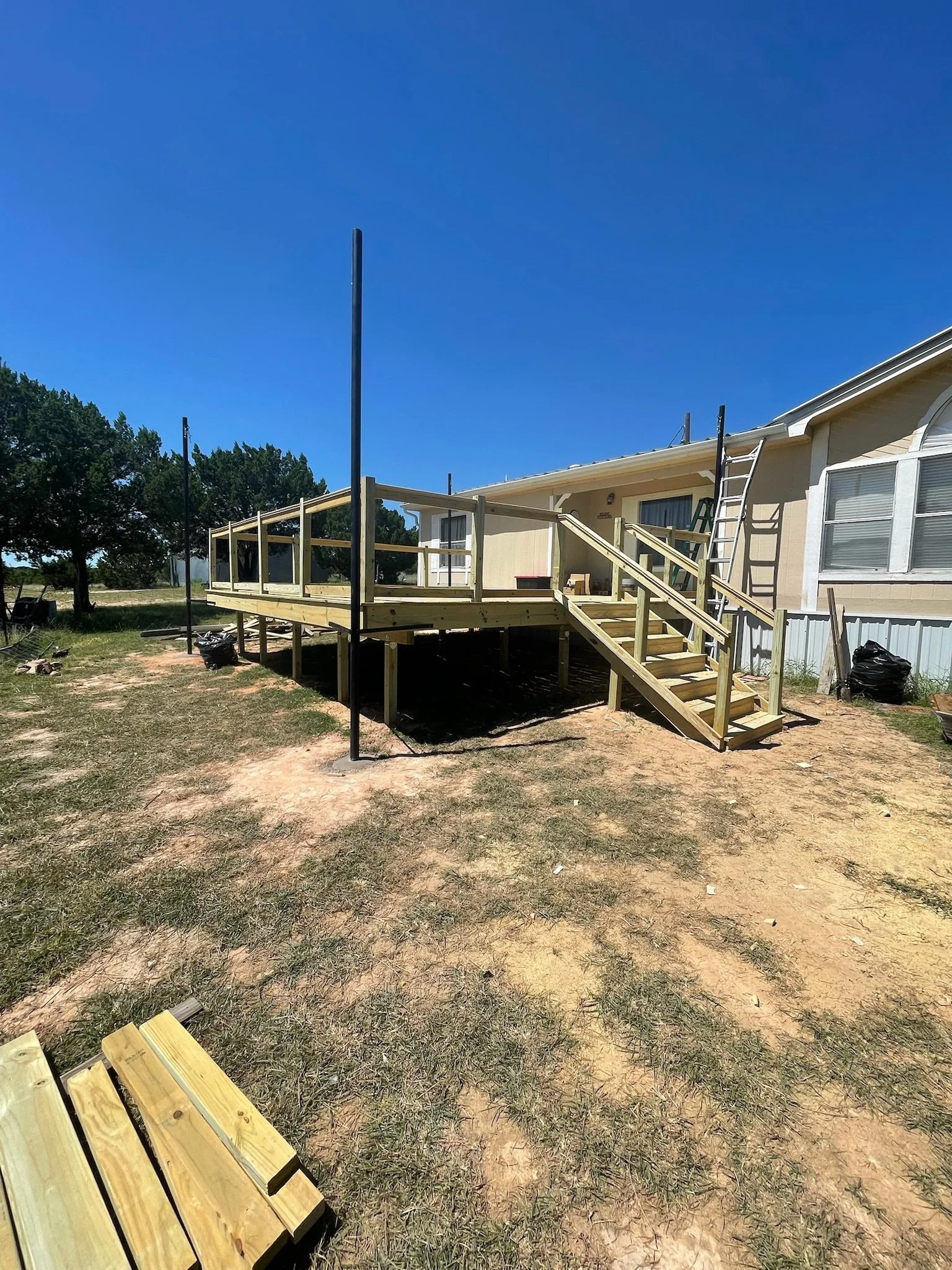 Construction of a wooden deck attached to the side of a house under clear blue sky, with building materials and tools nearby.