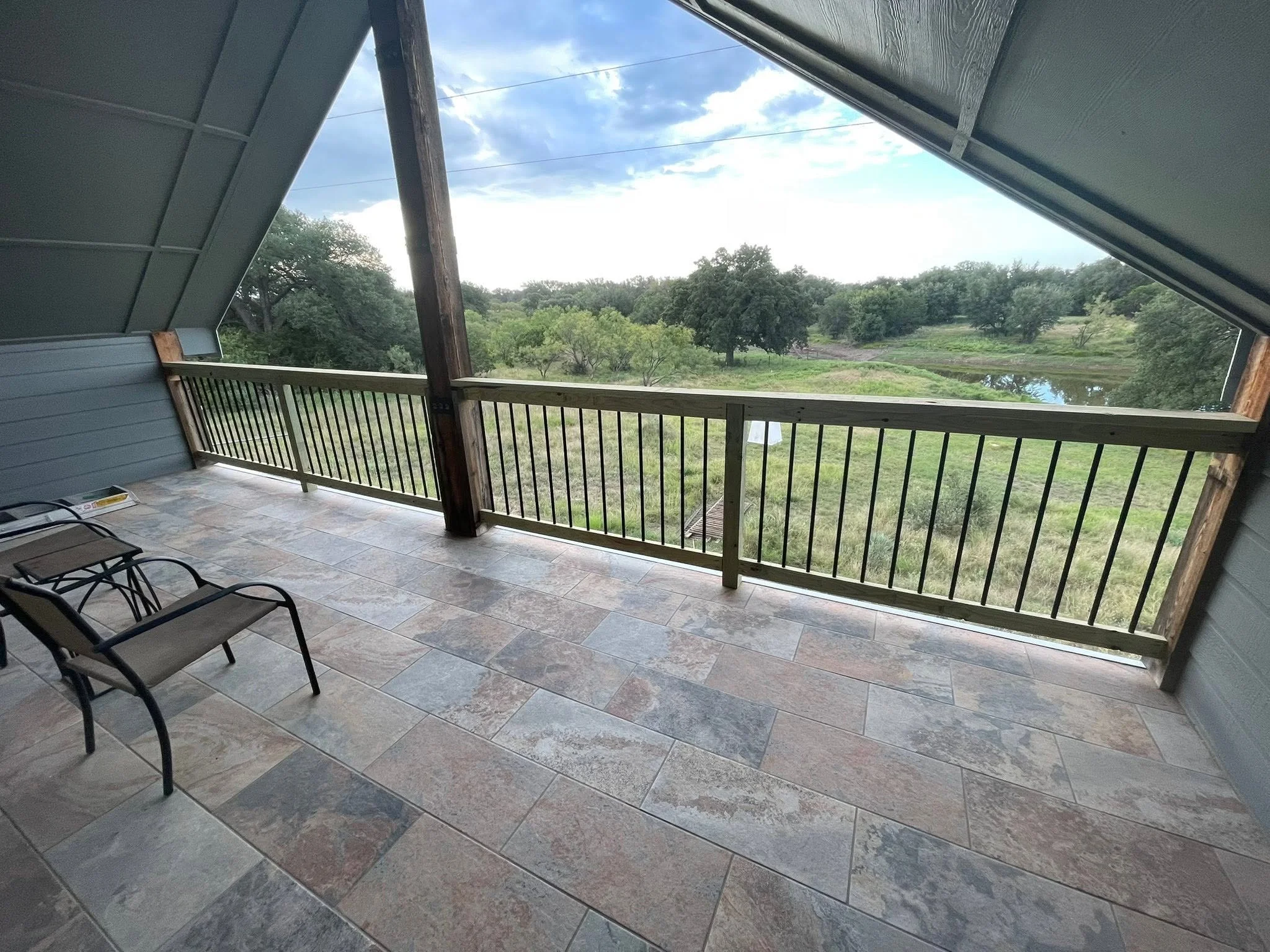 View from a covered porch or balcony overlooking a grassy area with trees, a pond, and a cloudy sky.