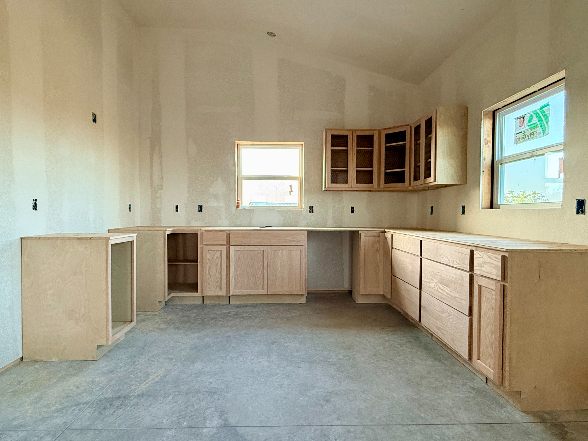 Interior of a room under construction with unfinished wooden cabinets and drawers, and three windows letting in natural light.