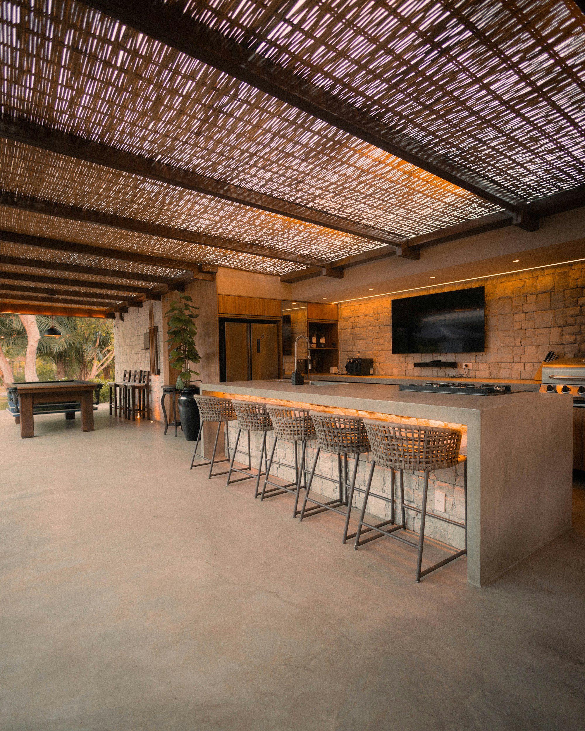 Modern indoor kitchen area with a large island, barstools, and exposed brick walls. A black flat screen TV is mounted on the wall, and there are wooden cabinets and a tall refrigerator.