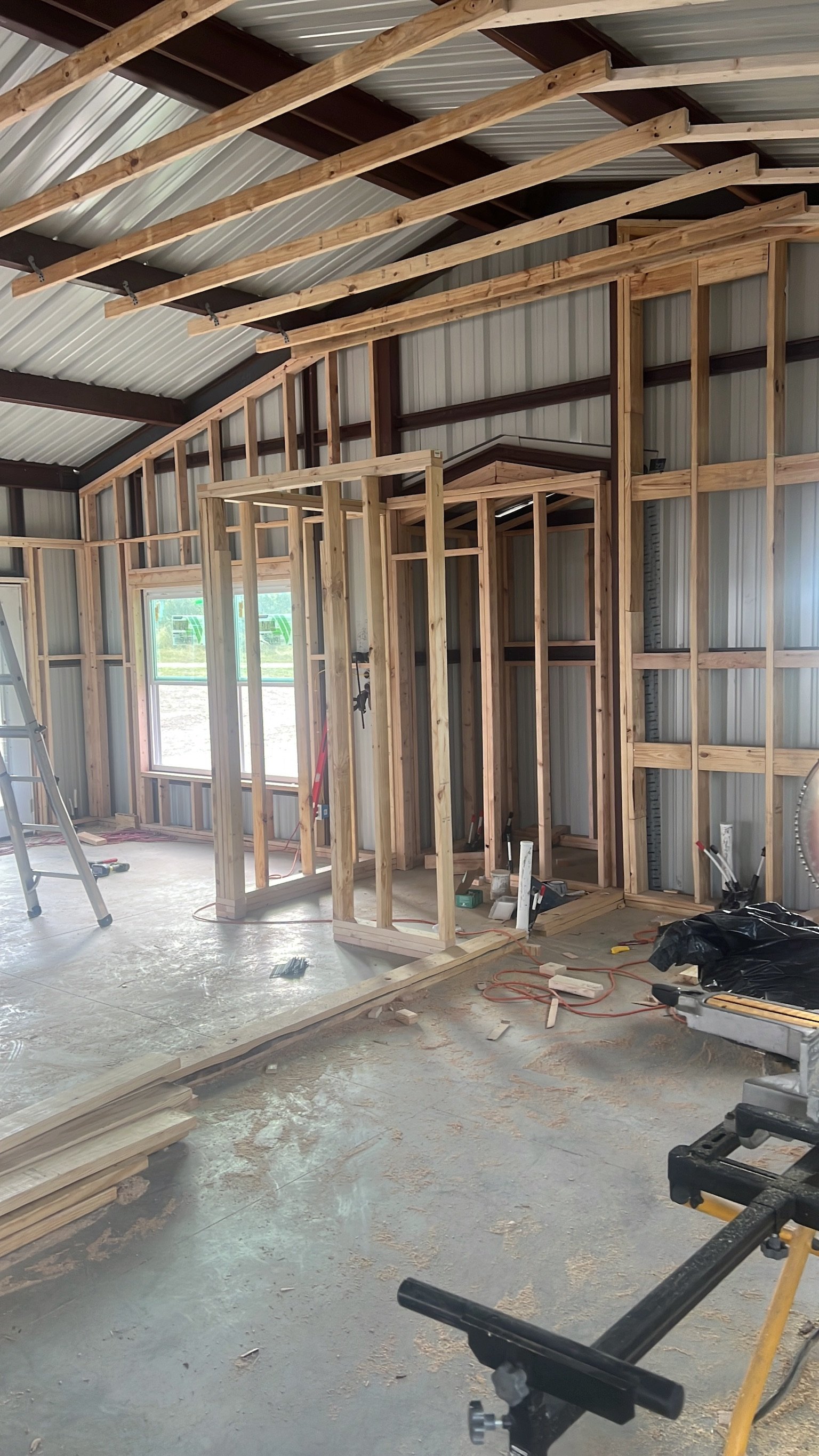 Interior view of a house under construction, with wooden framing for walls and ceiling, construction tools, and materials scattered on the floor.
