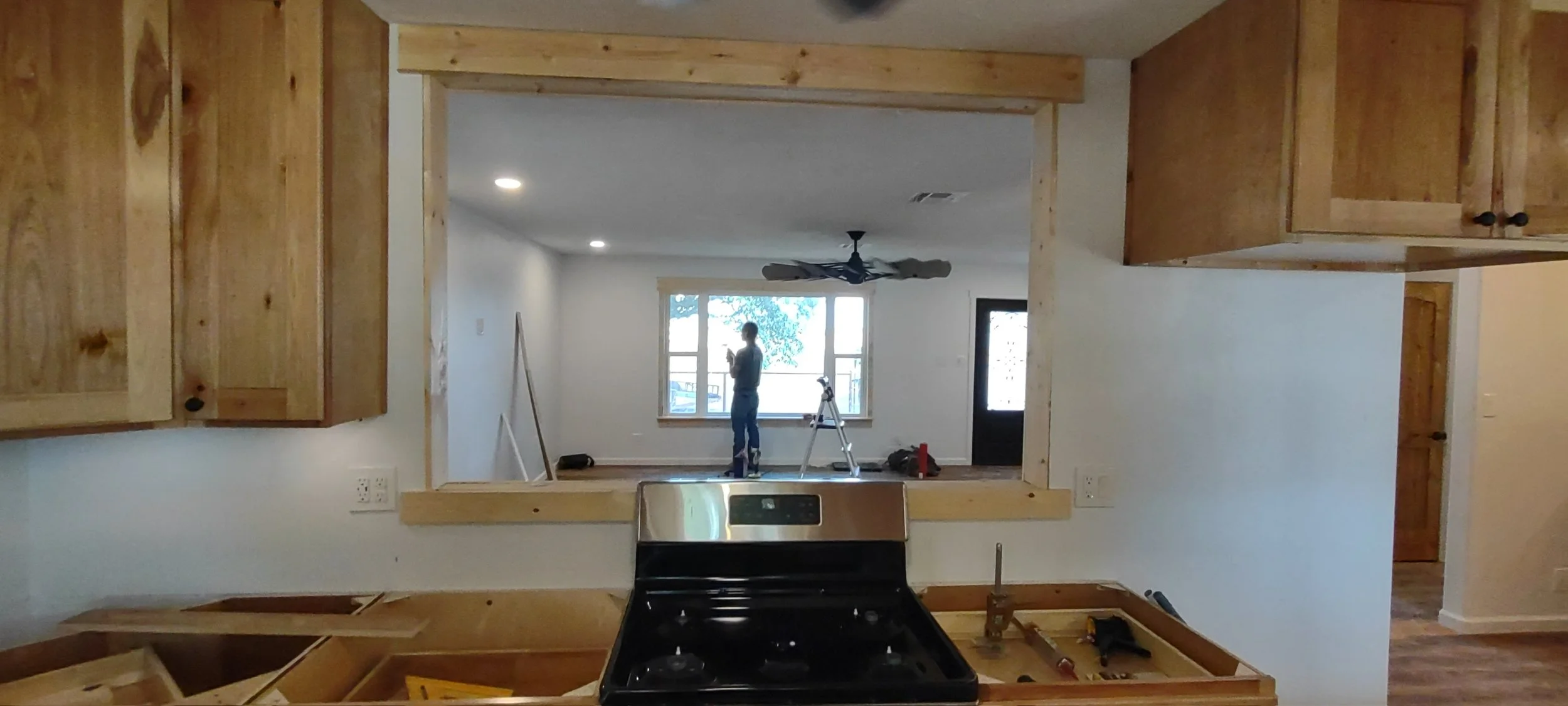 A person standing on a ladder inside a room, installing or working on a ceiling fan, with a window in the background. The photo is taken from the kitchen area, looking into the living space, with visible wooden cabinets and a stove in the foreground.