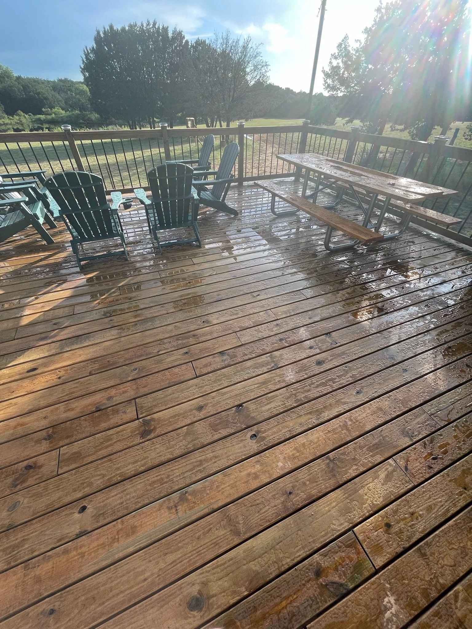 A wooden deck with wet surface, surrounded by a black metal fence. There are outdoor seating and a picnic table on the deck. The background features trees, grass, a blue sky with some clouds, and bright sunlight creating lens flare.