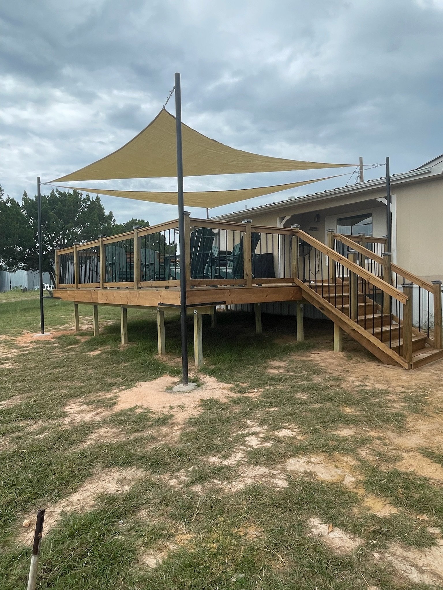 A wooden deck with stairs and black metal railings attached to a house. Two large beige shade sails are stretched above the deck, secured to tall black poles. There are outdoor chairs and a grill on the deck. The sky is cloudy, and there are trees in