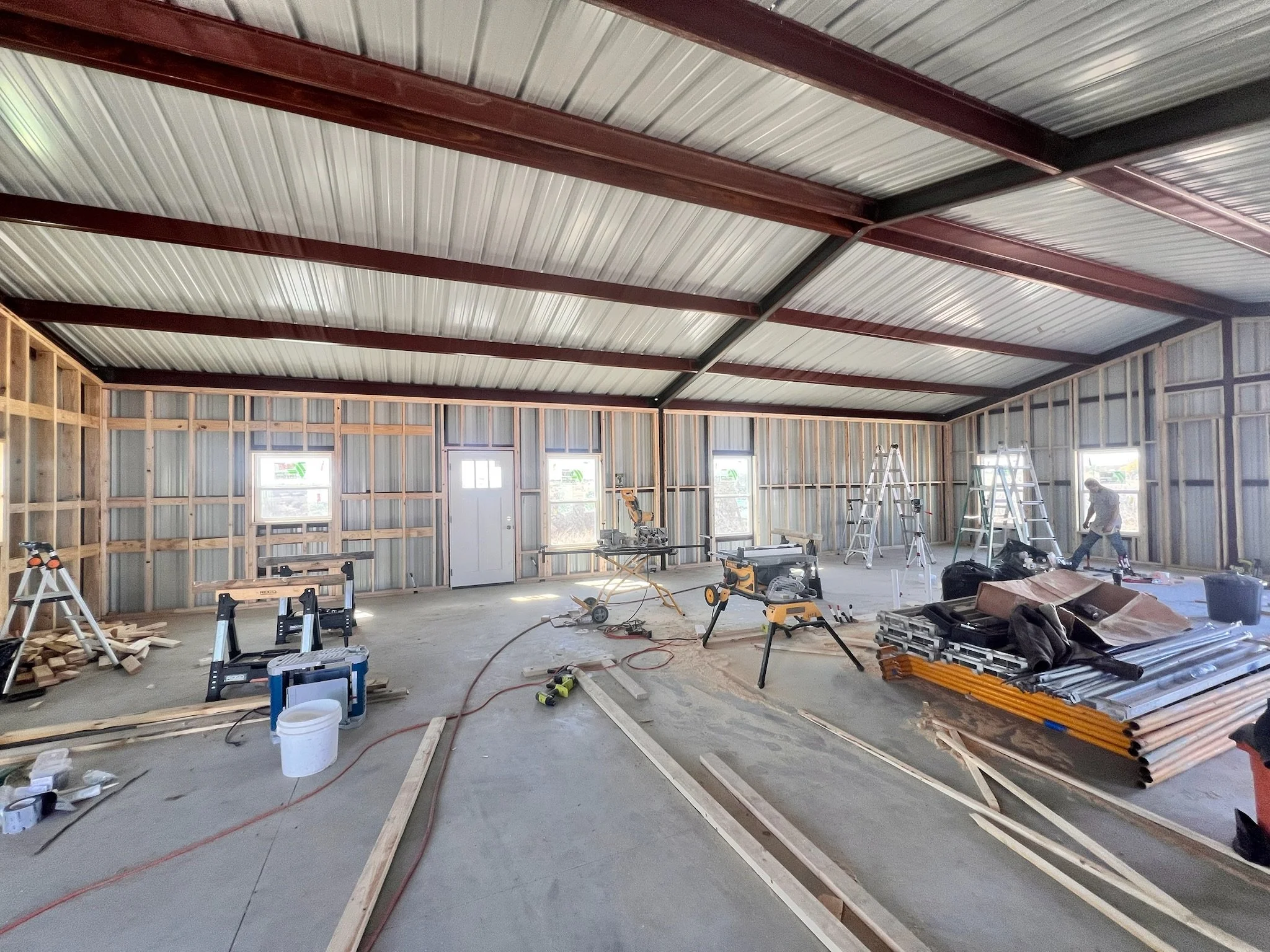 The interior of a building under construction with exposed wooden framing and a metal roof. Construction tools, ladders, and building materials are scattered throughout the space, with workers visible in the background.